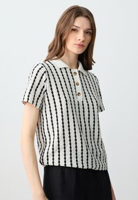 Polo en tricot crème avec des rayures noires verticales. Dispose d'un col et de trois boutons dorés pour la fermeture. Manches courtes, coupe décontractée.