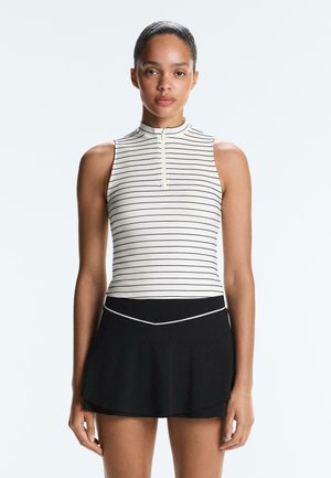 Žena s vlasmi stiahnutými dozadu, oblečená v bielom tielku bez rukávov s čiernymi pásmi a predným zipsom, v kombinácii s čiernou sukňou.