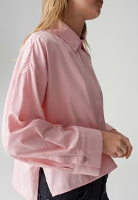 Button-up shirt in soft pink fabric, featuring a classic collar, loose fit, and cuffed sleeves. Notable for its clean design and seam details.