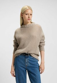 Beige gebreide trui met ribbelstructuur, ronde halslijn en opgerolde mouwen, gecombineerd met blauwe denim jeans en een zwarte riem.