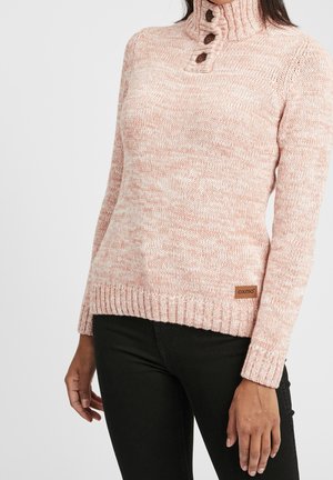 Frau trägt einen rosa melierten Strickrollkragenpullover mit braunen Knöpfen und schwarze Hose, steht vor einem schlichten hellen Hintergrund.