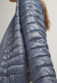 Personne aux cheveux longs porte une veste matelassée bleue brillante, vue de côté avec le bras tendu, en intérieur devant un fond uni.
