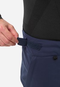 Bretelle ajustable bleu marine avec fermeture Velcro, attachée à un vêtement en tissu léger et texturé, montrant des détails de couture.