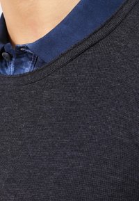 Närbild av en marinblå stickad tröja lager på lager över kragen på en blå skjorta som sitter runt en persons hals och övre bröst.