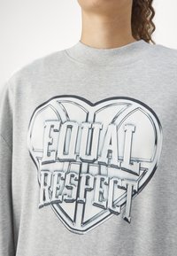Personne portant un sweat-shirt gris avec un grand motif en forme de cœur et les mots "Respect Égal" imprimés sur le devant.