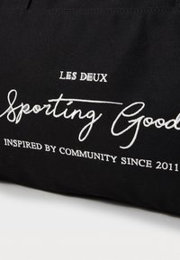 Tote de tela negra con texto bordado en blanco: "LES DEUX Sporting Goods" y "INSPIRADO POR LA COMUNIDAD DESDE 2011". Diseño simple, material duradero.