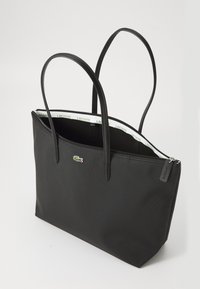 Sac fourre-tout Lacoste noir avec deux poignées, ouverture par zip en haut et petit logo crocodile à l'avant. Bord intérieur estampillé du texte "Lacoste".