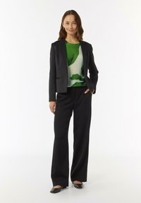 Zwarte op maat gemaakte blazer over een groen top met een bladpatroon, gecombineerd met zwarte wijde broek en zwarte flats.
