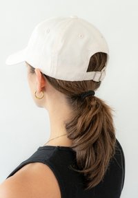 Gorra de béisbol de color rosa claro con visera curva, correa ajustable en la parte posterior y una marca sutil. Cabello recogido en una coleta, usando pendientes de aro.