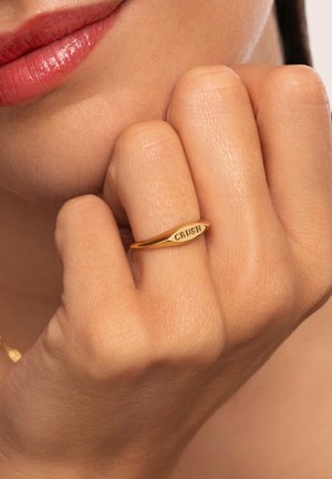 Goldring mit ovaler Form, graviert mit dem Wort "CRUSH." Die Hand zeigt eine natürliche Hautstruktur und berührt leicht das Gesicht.