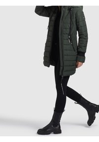 Zielona, pikowana kurtka puchowa z zamkiem błyskawicznym i regulowanym kapturem, zestawiona z czarnymi spodniami typu skinny i czarnymi botkami za kostkę.