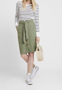 Gonna di lino verde con vita legata e bottoni davanti, abbinata a una maglietta a righe a manica lunga e scarpe da ginnastica bianche alte. Borsa intrecciata tenuta in mano.