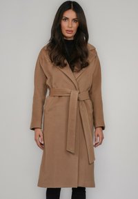 Beżowy wełniany płaszcz z paskiem, długimi rękawami, dużymi klapami i bocznymi kieszeniami; prezentowany w prostej, sięgającej do połowy długości fasonie.