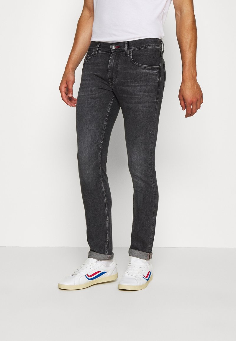 Man som bär slim-fit mörkgrå jeans med uppvikta benslut och vita sneakers med blå och röda accenter mot vit bakgrund.