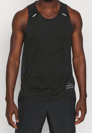 Mann trägt ein schwarzes ärmelloses Sporttanktop und schwarze Shorts, steht vor einem schlichten hellen Hintergrund.