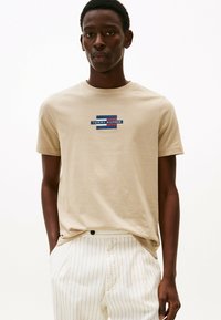 Camiseta de algodón beige con cuello redondo y mangas cortas, que presenta un gráfico del logo en azul, rojo y blanco centrado en el pecho.