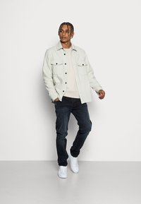 Hellegrau knöpfbares Hemd mit zwei Brusttaschen, kombiniert mit einem beigen Oberteil und dunklen Jeans, abgerundet mit weißen Turnschuhen. Einheitlicher Hintergrund.