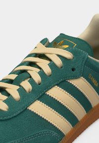 Πράσινο αθλητικό παπούτσι Adidas Samba με μπεζ ρίγες, μπεζ κορδόνια και σόλα από καουτσούκ, με το λογότυπο της μάρκας και την επιγραφή "SAMBA" στο πλάι.