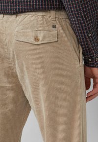 Beige Cordhosen mit vertikalen Rippen, ausgestattet mit einer Gesäßtasche, die mit einem Knopf gesichert ist. Der Stoff wirkt weich und strukturiert.