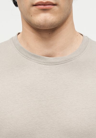 Κοντινό πλάνο του κάτω μέρους του προσώπου και του λαιμού ενός ατόμου που φοράει ένα ανοιχτό μπεζ μπλουζάκι με στρογγυλή λαιμόκοψη σε απλό φόντο.