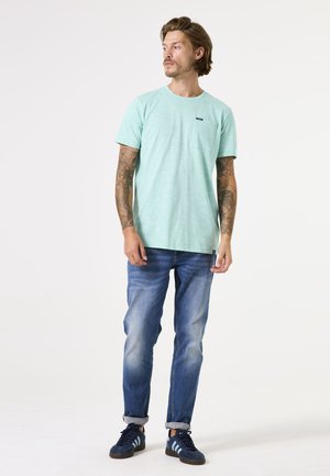 Light green short-sleeve t-shirt with a chest pocket, worn with rolled jeans and navy sneakers. Features a textured fabric and casual fit.