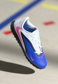 Λευκό και μπλε ποδοσφαιρικό παπούτσι Nike με ροζ λογότυπο swoosh που αιωρείται πάνω από το δάπεδο του γηπέδου με χρωματιστές γραμμές και σήματα.