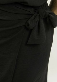 Jupe noire avec un détail de nœud noué à la taille, fabriquée en tissu doux avec une texture lisse et un design légèrement drapé.