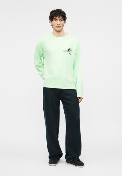 Άνδρας με κατσαρά μαλλιά που φοράει ανοιχτοπράσινο μακρυμάνικο πουκάμισο με γραφικό στο στήθος, φαρδιά μαύρη παντελόνια και μαύρα αθλητικά παπούτσια, στέκεται μπροστά σε λευκό φόντο.