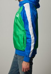 Grön och blå huva jacka med vita sidostreck och dragkedja. Har en glänsande, slät textur och elastiska manschetter.