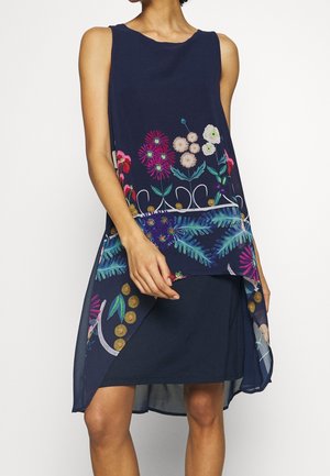 Ärmelloses marineblaues Kleid mit mehrlagigem Blumenmuster-Overlay, das bunte Blumen und grüne Blätter zeigt, getragen von einer stehenden Person.