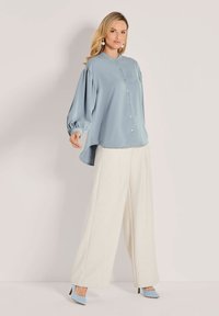 Eine Frau trägt eine lockere, hellblaue Bluse mit Knöpfen, weite weiße Hosen, hellblaue Absatzschuhe und Perlenohrringe. Sie steht vor einem schlichten Hintergrund.