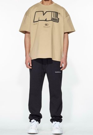 Beiges übergroßes T-Shirt mit schwarzem Grafikdruck, getragen zu schwarzen Jogginghosen mit weißem Logodetail, kombiniert mit weißen Sneakern.