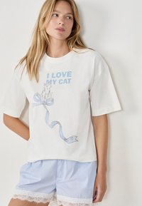 Mujer vistiendo una camiseta blanca con el texto "AMO A MI GATO" y una ilustración de gato, combinada con pantalones cortos a rayas azules con borde de encaje.