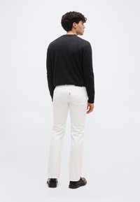 Άνδρας που φοράει σκούρο πουκάμισο με μακριά μανίκια, λευκά τζιν και μαύρα παπούτσια, στέκεται με την πλάτη σε έναν απλό φωτεινό φόντο.