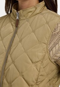 Mönstrad beige väst med hög krage, som har en framtill dragkedja och en subtil logotyp-mönster, kombinerad med en strukturerad stickad tröja under.