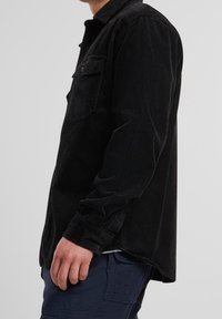 Chemise en velours côtelé noir avec une fermeture à boutons sur le devant, une poche poitrine unique et des manches longues, présentant une texture côtelée subtile.