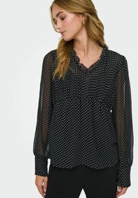 Femme portant un chemisier noir transparent à pois blancs, manches longues bouffantes et encolure à volants, associé à un pantalon noir.