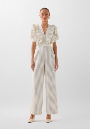 Jumpsuit bianco a righe con maniche corte arricciate, scollatura a V profonda e gambe larghe. Tessuto morbido con righe verticali a contrasto. Le scarpe sono beige.