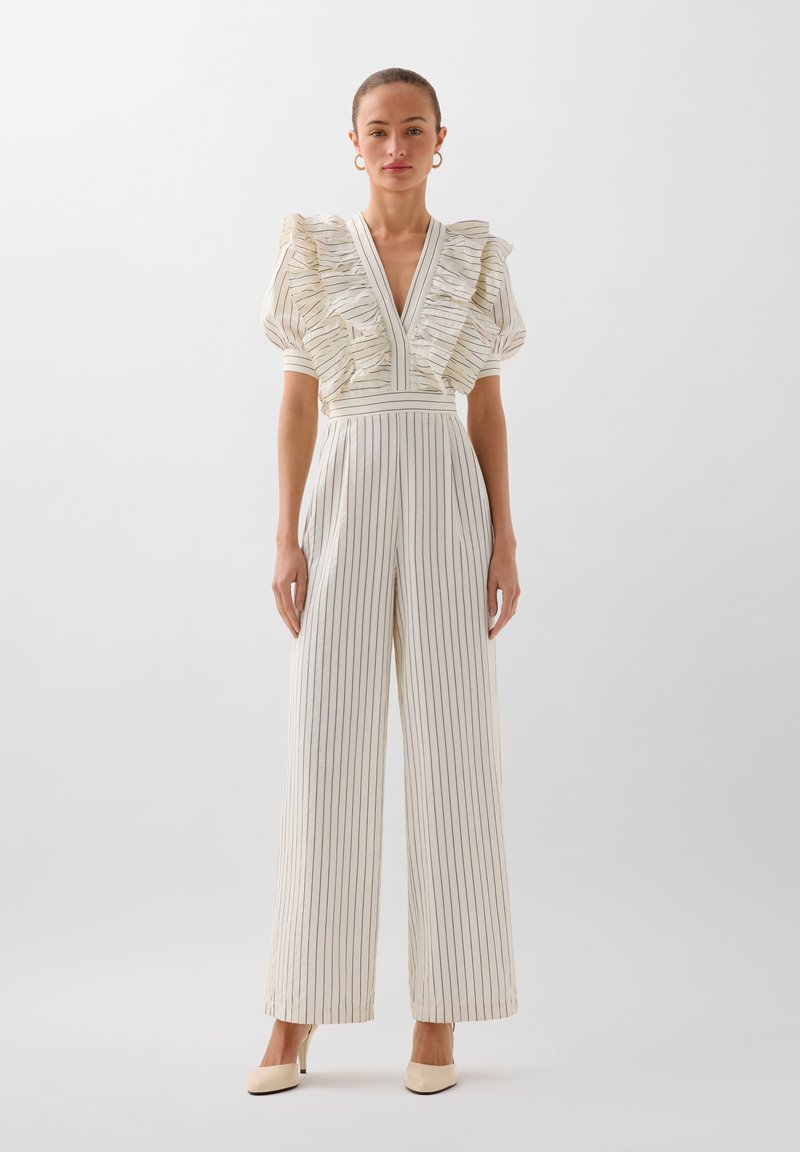 Jumpsuit bianco a righe con maniche corte arricciate, scollatura a V profonda e gambe larghe. Tessuto morbido con righe verticali a contrasto. Le scarpe sono beige.