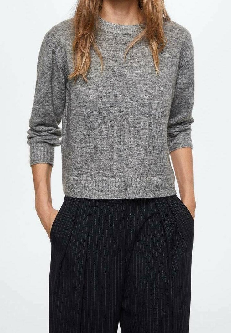 Personne portant un pull gris à manches longues avec les manches remontées et un pantalon noir à fines rayures, les mains dans les poches.