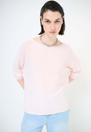 Donna con capelli corti scuri effetto bagnato, che indossa una camicia rosa chiaro, una collana a catena spessa color argento e jeans azzurri su sfondo bianco.