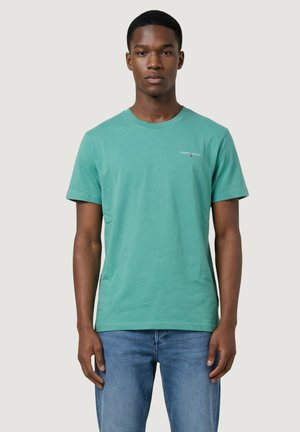 Joven con una camiseta azul verdosa de Tommy Jeans y vaqueros azules, de pie contra un fondo claro y liso, mirando hacia adelante.