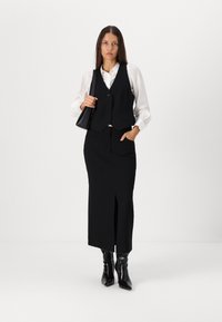 Black tailored vest over a white shirt, paired with a black pencil skirt featuring a side slit, and black heeled boots. Holds a small black bag.
