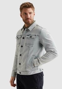 Muž s bradou v svetlosivej džínsovej bunde na gombíky, s jednou rukou v vrecku, pozerajúci sa bokom na jednoduchom pozadí.