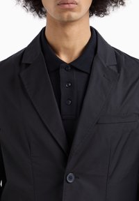 Blazer nero con una polo nera a trama sotto. Le caratteristiche includono una chiusura a un solo bottone, revers e una tasca frontale.