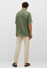 Zielona koszula z krótkim rękawem wykonana z teksturowanej tkaniny, w zestawie z jasnobeżowymi spodniami i sandałami, widziana od tyłu na jednolitym tle.