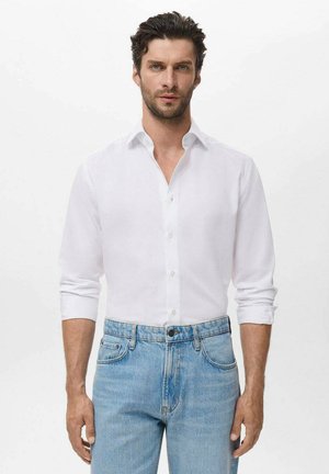 Hombre con el cabello rizado y corto de color oscuro lleva una camisa blanca de botones con las mangas remangadas y jeans de color azul claro, de pie contra un fondo sencillo.