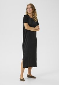 Black, short-sleeve, midi-length dress made of smooth fabric with side slits and a rounded neckline. Featured with flat slip-on shoes.