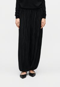 Άτομο που φοράει μαύρο πλισέ παντελόνι με φαρδιά μπατζάκια και μαύρα μυτερά φλατ παπούτσια, στέκεται μπροστά σε απλό λευκό φόντο.