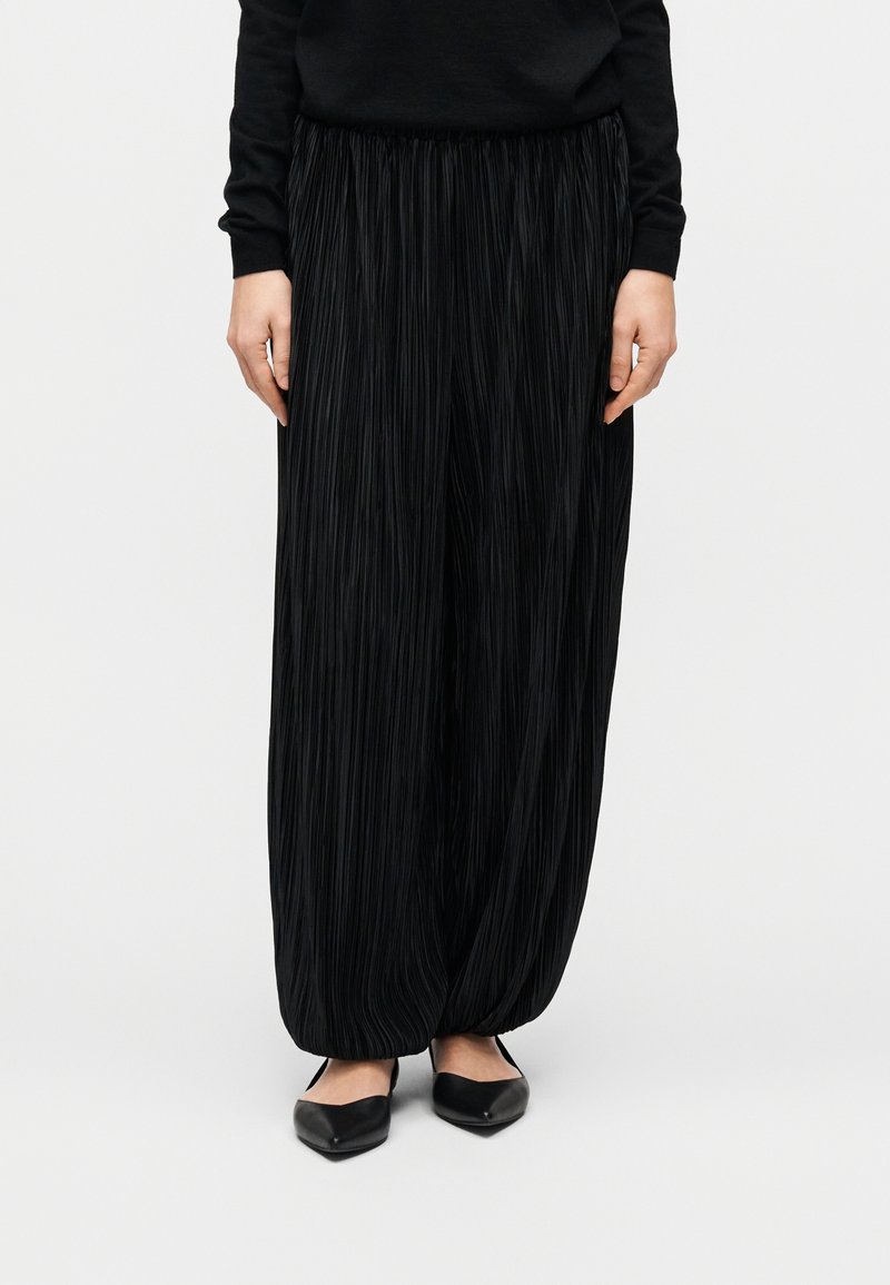 Άτομο που φοράει μαύρο πλισέ παντελόνι με φαρδιά μπατζάκια και μαύρα μυτερά φλατ παπούτσια, στέκεται μπροστά σε απλό λευκό φόντο.